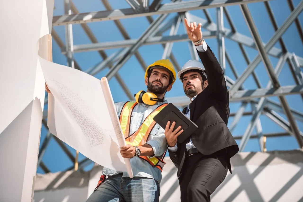 An engineer holding a blueprint and an architect in a suit pointing upward while inspecting a steel roof frame at a construction site.
