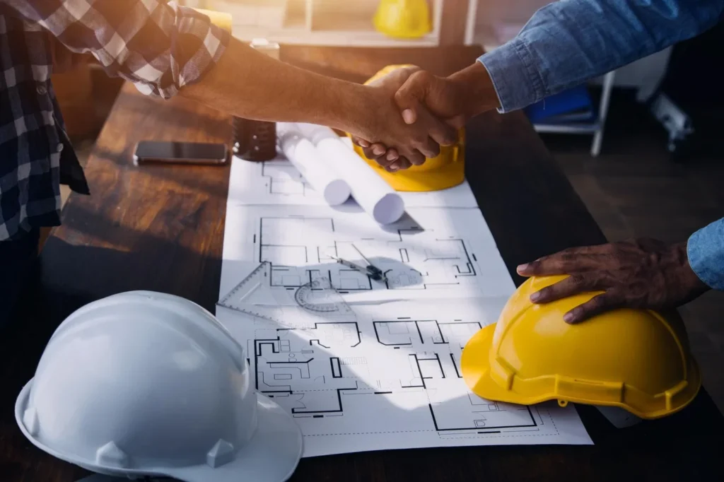 Close-up of construction managers shaking hands over architectural blueprints and hardhats, representing successful project consultation