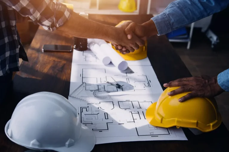 Close-up of construction managers shaking hands over architectural blueprints and hardhats, representing successful project consultation